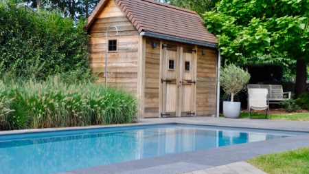landelijk tuinhuis staat naast een lichtgrijs zwembad met rolluikplage en trap in een landelijke tuin
