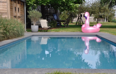 roze flamingo ligt in het gras naast een lichtgrijs zwembad met rolluikplage en trap